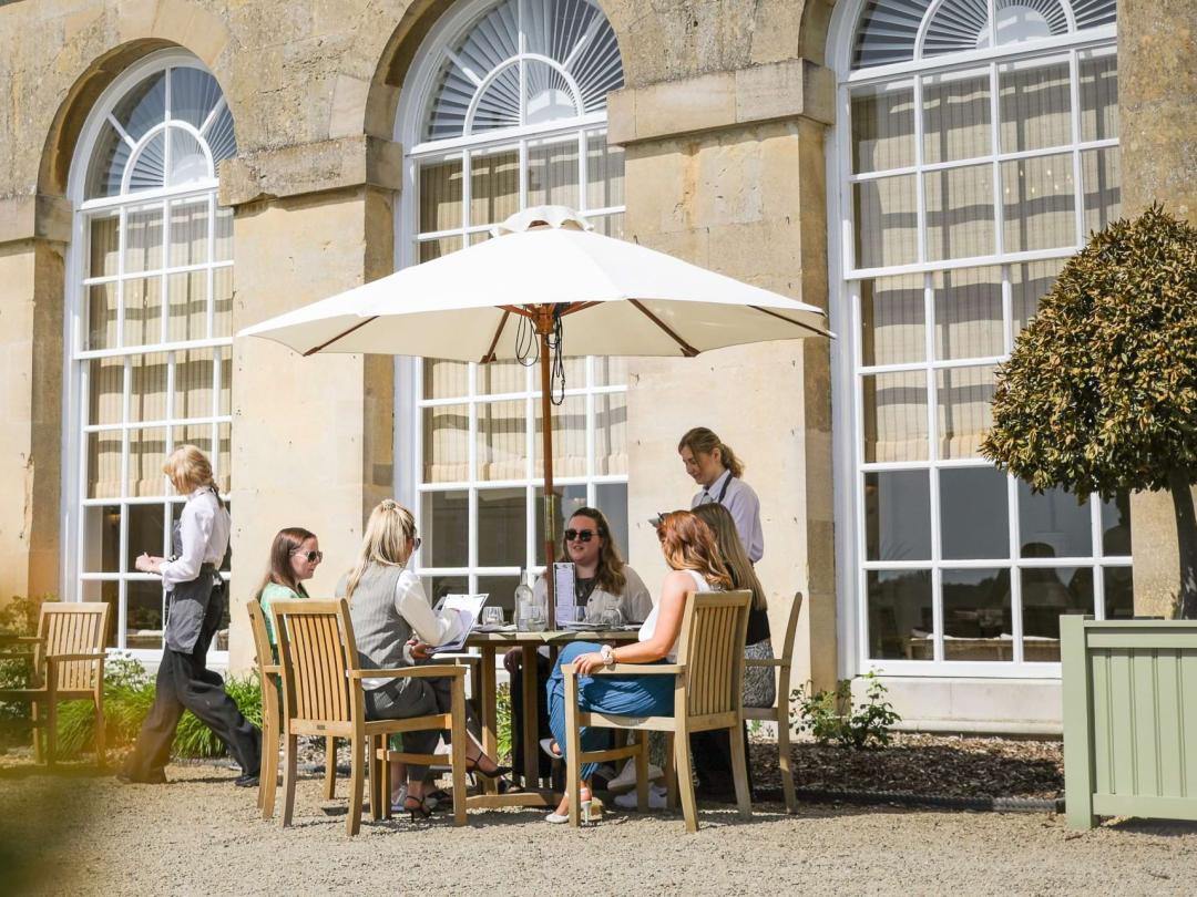 Guests dining at an outdoor table under a parasol beside the arched windows of the Orangery