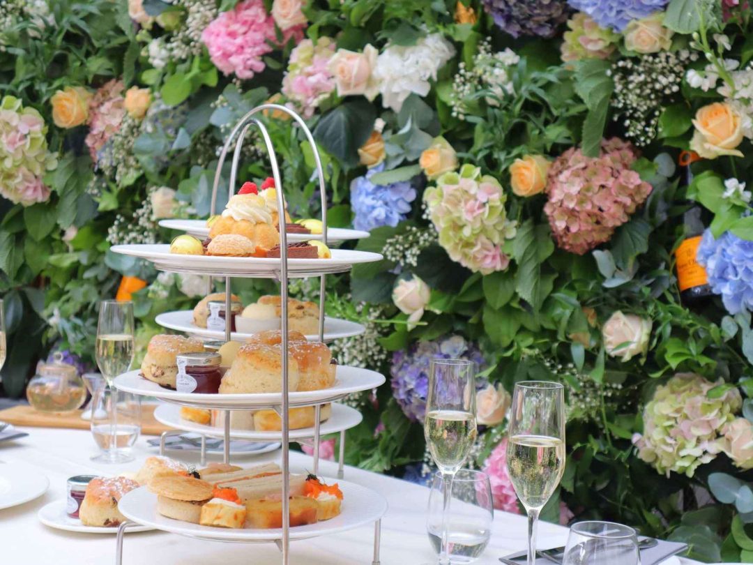 A three-tier afternoon tea stand of scones and cakes beside glasses of sparkling wine, against a floral display