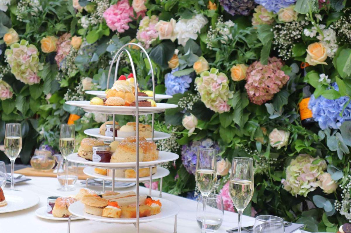 A three-tier afternoon tea stand of scones and cakes beside glasses of sparkling wine, against a floral display
