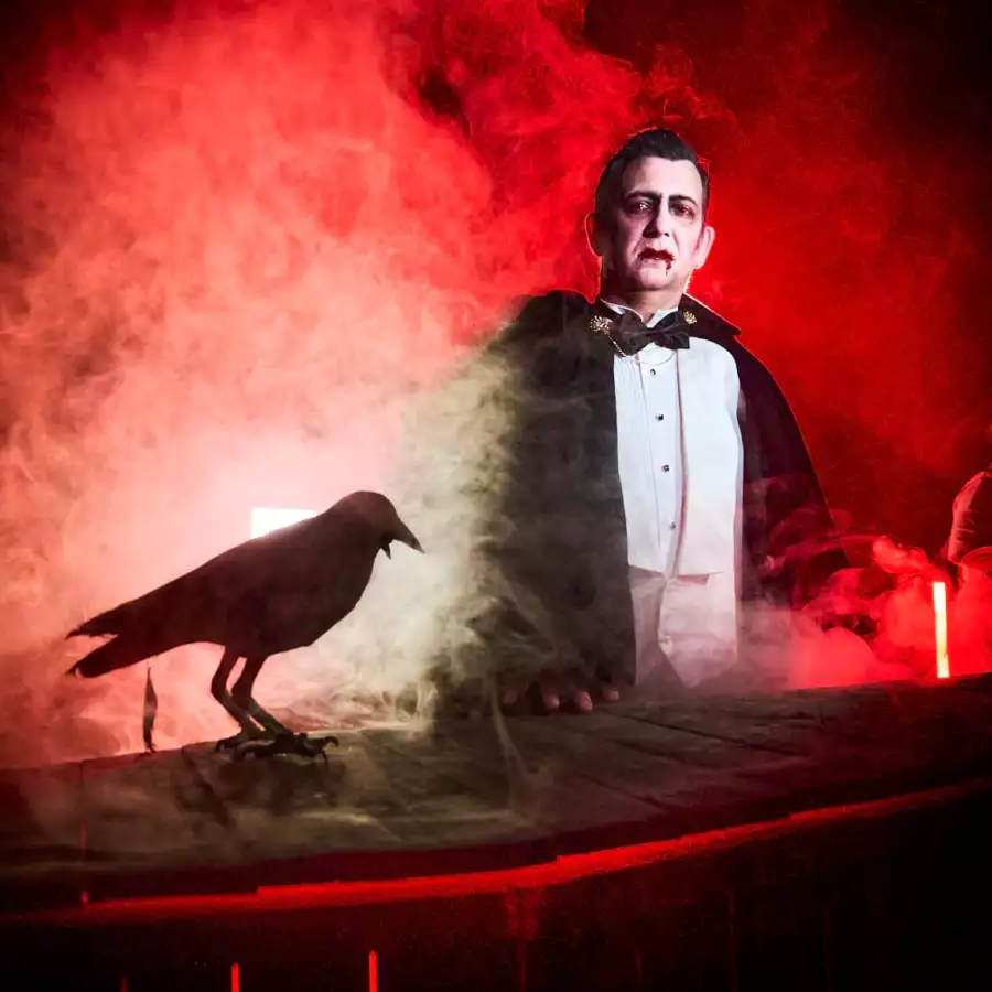 Actor dressed as a vampire in a black cape and bow tie stands in red smoke beside a perched raven