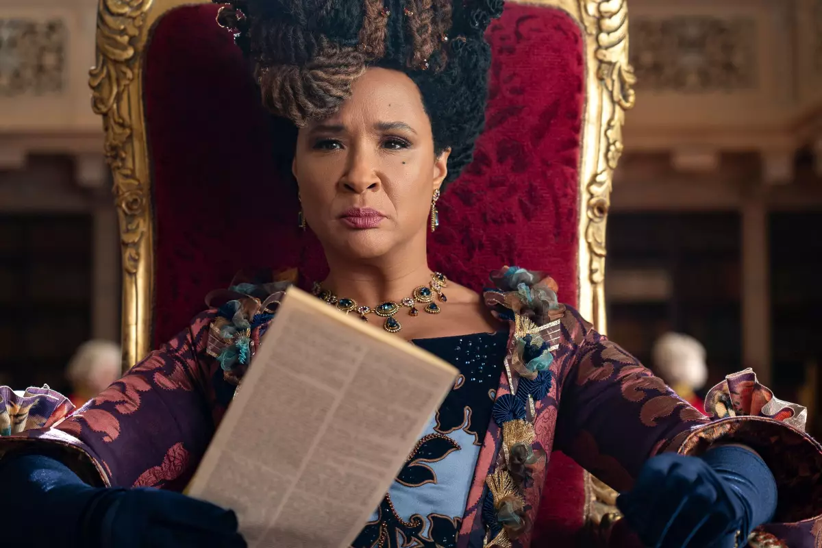 A woman in an ornate patterned gown and elaborate hairstyle sits on a red and gold throne holding a newspaper, in a still from a filmed drama