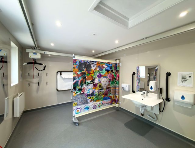 A Changing Places accessible toilet with a ceiling hoist, height-adjustable changing bench and a colourful printed screen