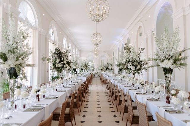 A long arched hall set for a wedding meal, with banquet tables, chandeliers and tall white flower arrangements