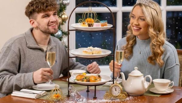 A couple toasting with sparkling wine over a Christmas afternoon tea decorated with a glass slipper and golden carriages, the woman wearing a tiara