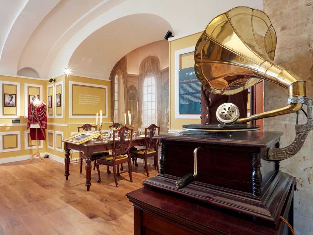 An exhibition room with a brass horn gramophone, a laid dining table and a ceremonial robe on display