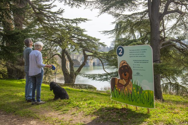 Two visitors and their dog beside the lake, next to a children's trail sign