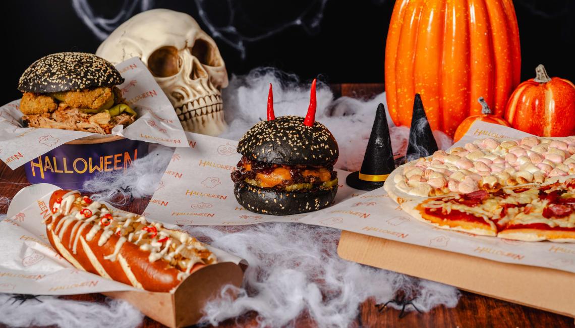 Halloween-themed burgers, a hot dog and pizza displayed with pumpkins and a skull