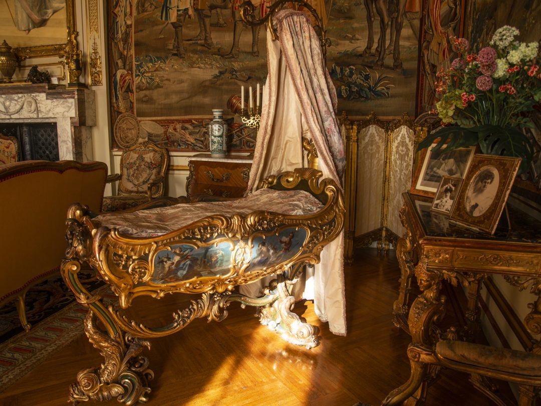 An ornate gilded cradle beneath a draped canopy in a tapestry-hung room