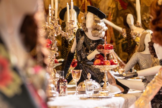 Mannequins in period costume seated at a candlelit table in a fashion exhibition