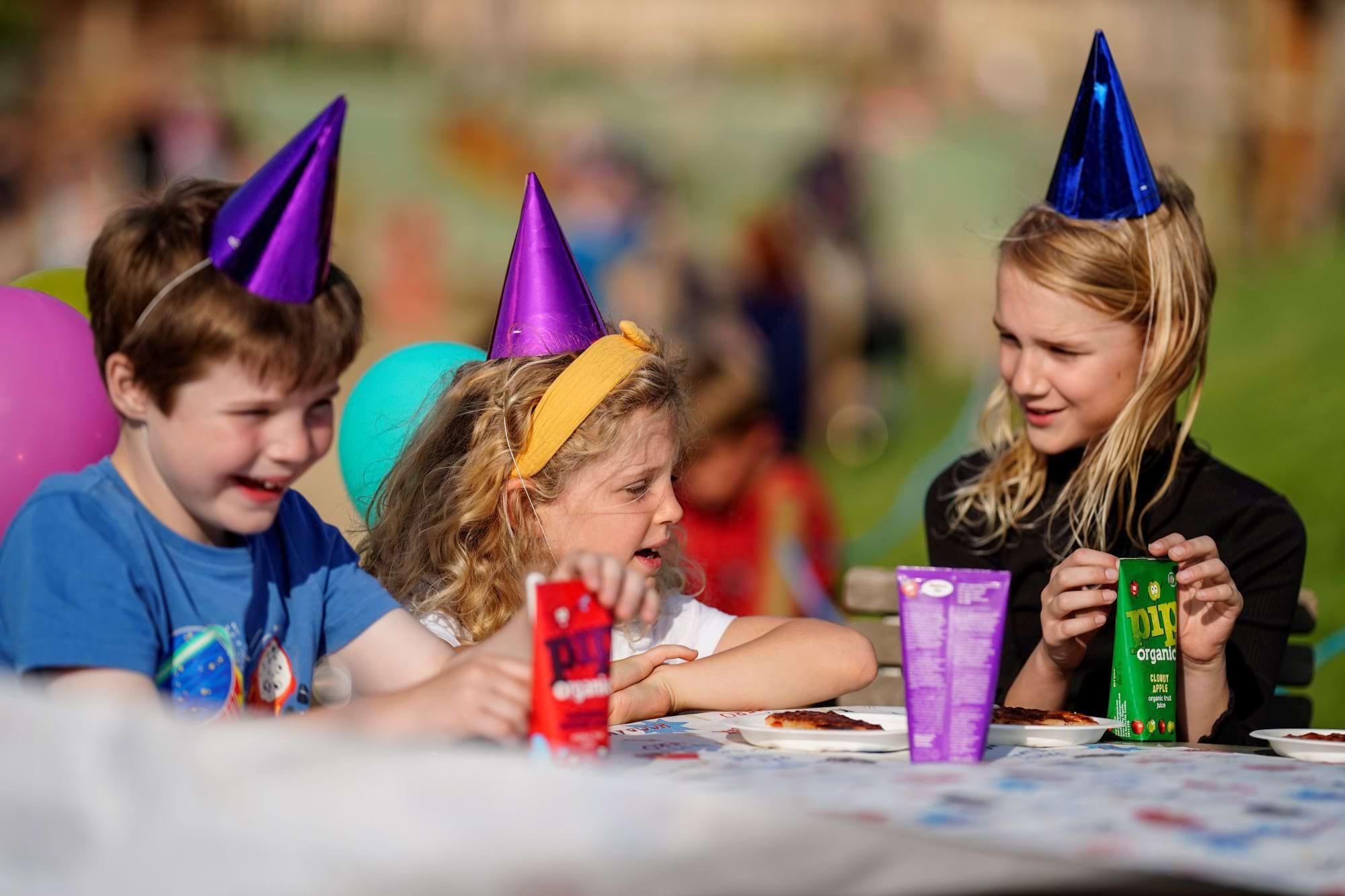 Three children in party hats eating pizza and drinking juice at an outdoor party table