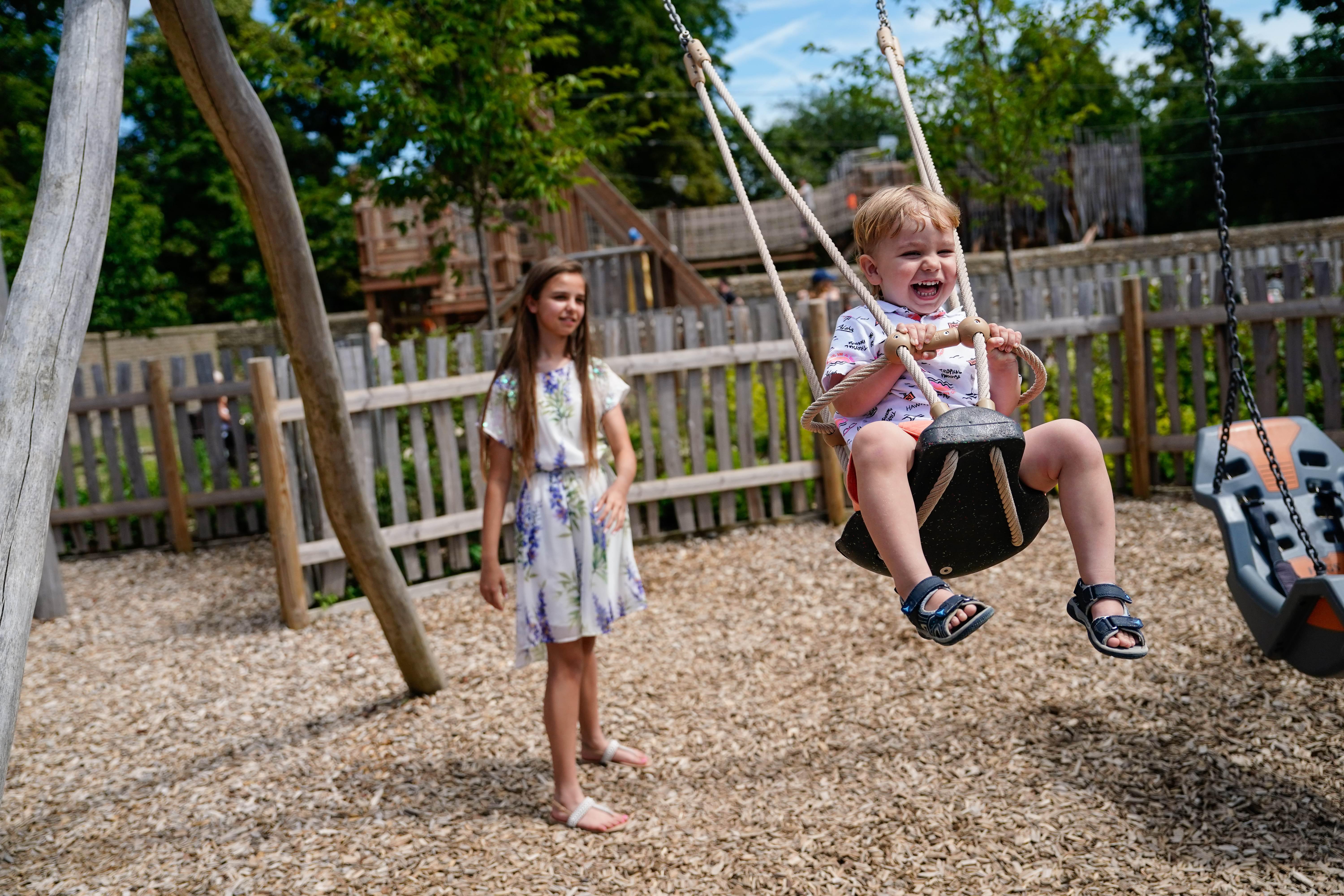 A toddler laughing on a rope swing in the adventure playground while an older girl watches