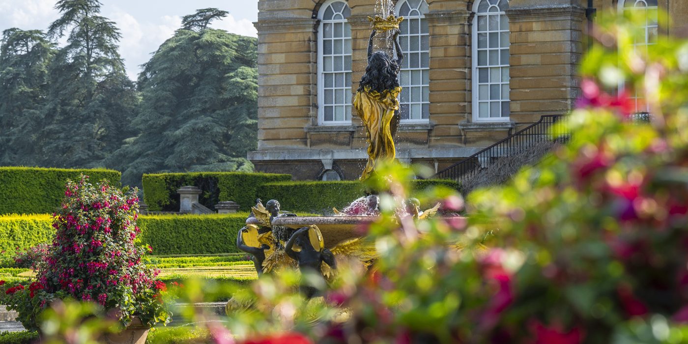 A gilded statue and fountain in the Italian Garden with pink flowers in the foreground and the Palace behind