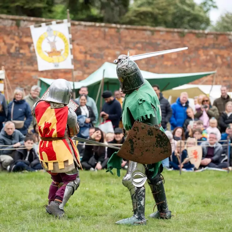 Two costumed fighters in armour duel on grass with swords as spectators watch in front of a brick wall