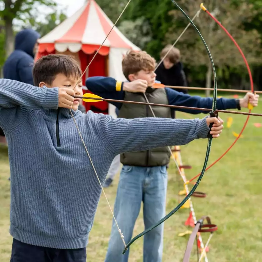 Two children draw bows at an archery stall at a medieval-themed event, with a red and white tent behind