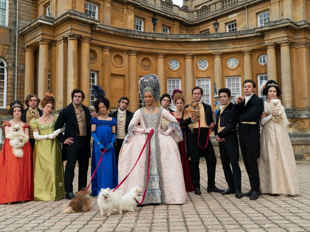 The cast of Queen Charlotte in Regency costume, with Pomeranian dogs on leads, posed in the Great Court of Blenheim Palace