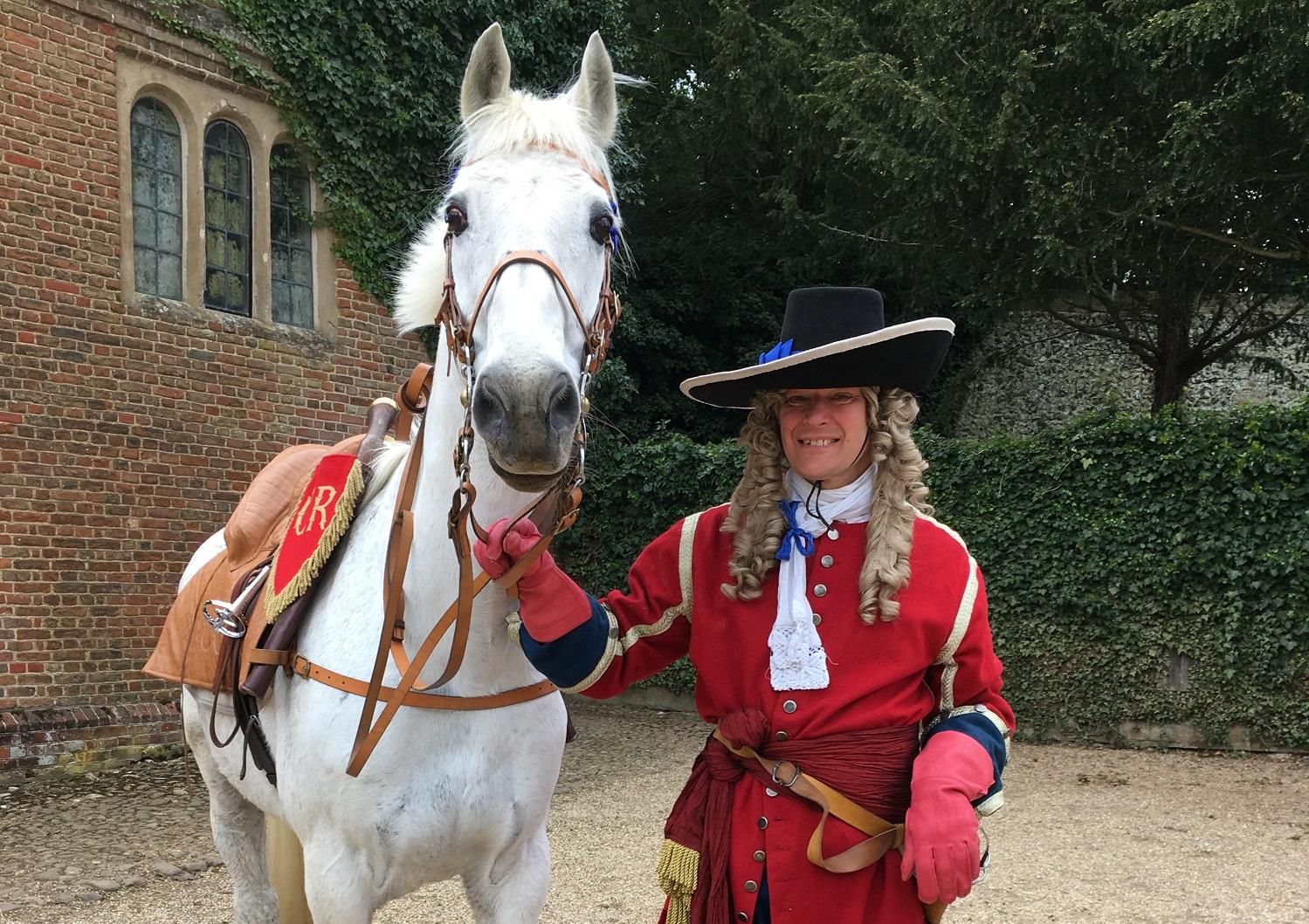A costumed interpreter in a red military coat and wig standing beside a white horse