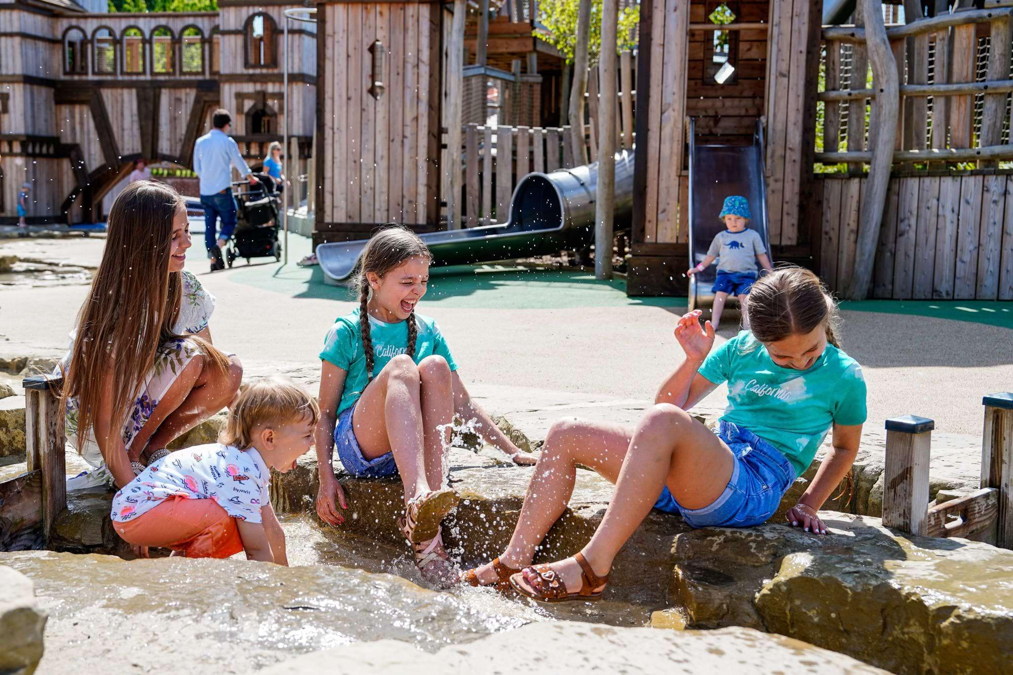 Children splashing in the water play stream in front of the wooden play fort at Adventure Play