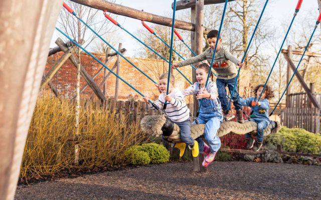 A toddler laughing on a rope swing in the adventure playground while an older girl watches