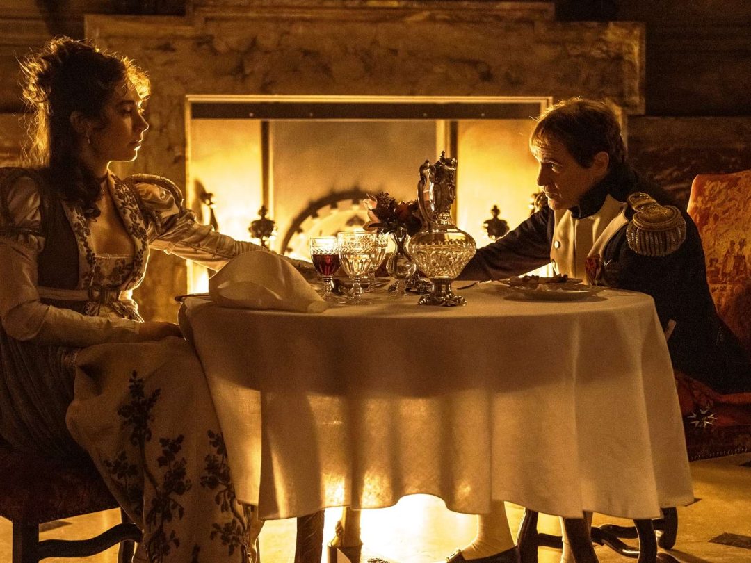 Two actors in period costume dining by candlelight at a small table in front of a fireplace, in a scene from the film Napoleon