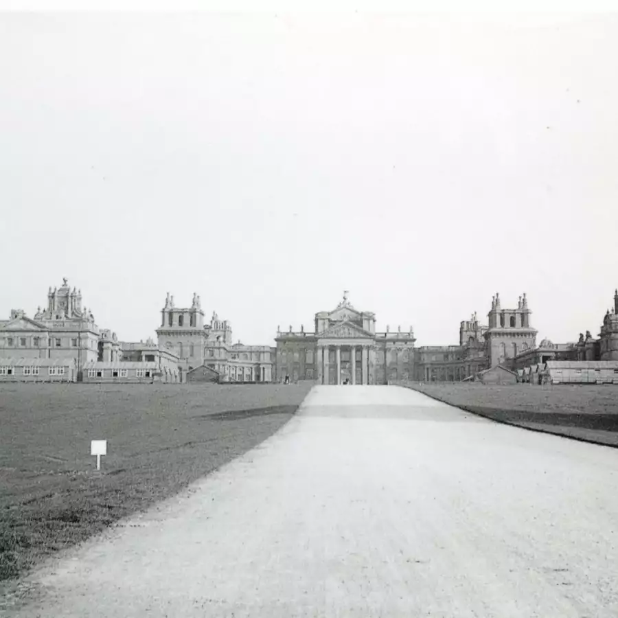Black and white historic view of the Palace across a wide gravel drive, with a small sign on the grass