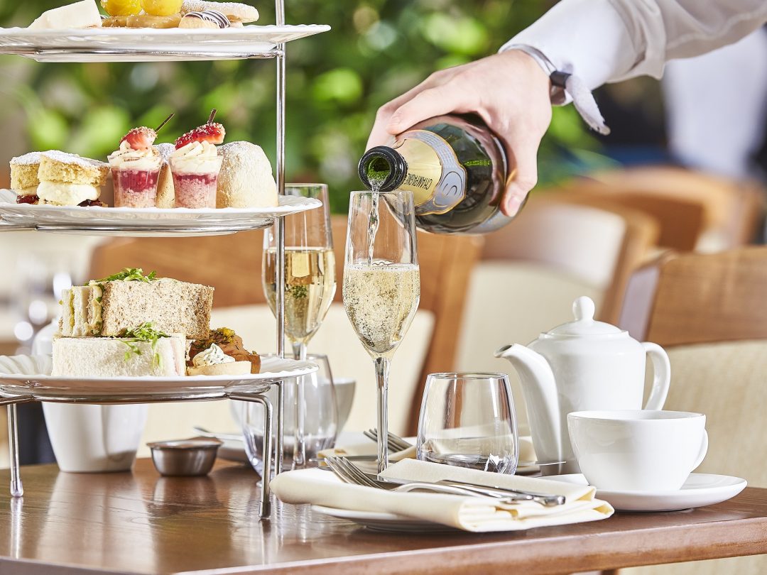 Sparkling wine being poured beside a tiered afternoon tea stand of sandwiches and cakes