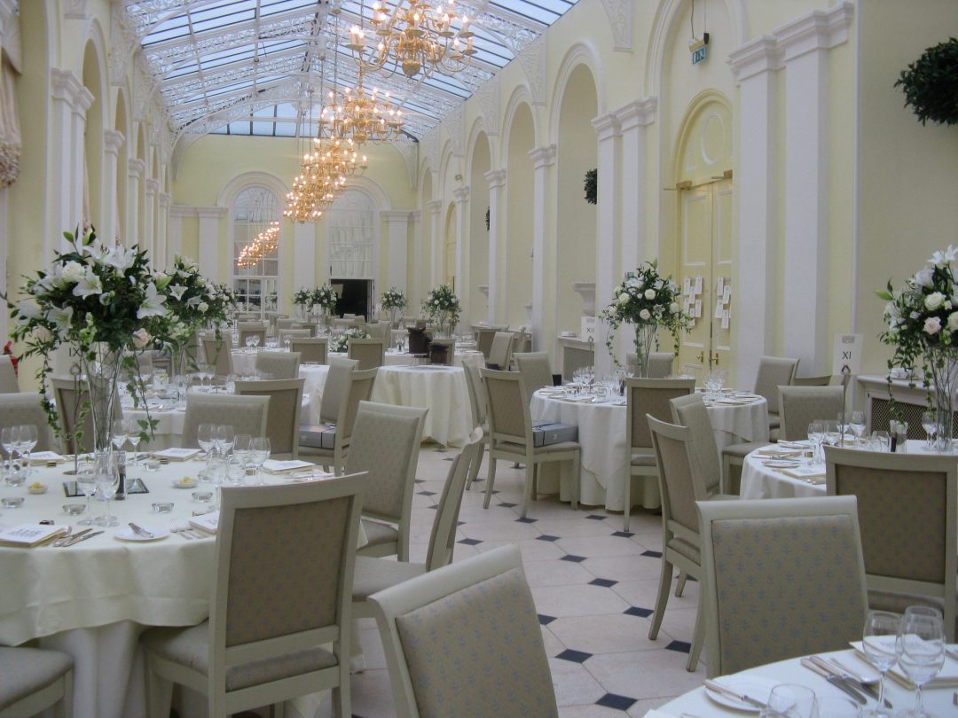 The Orangery laid for a formal dinner, with white linen and tall floral centrepieces beneath its glass roof