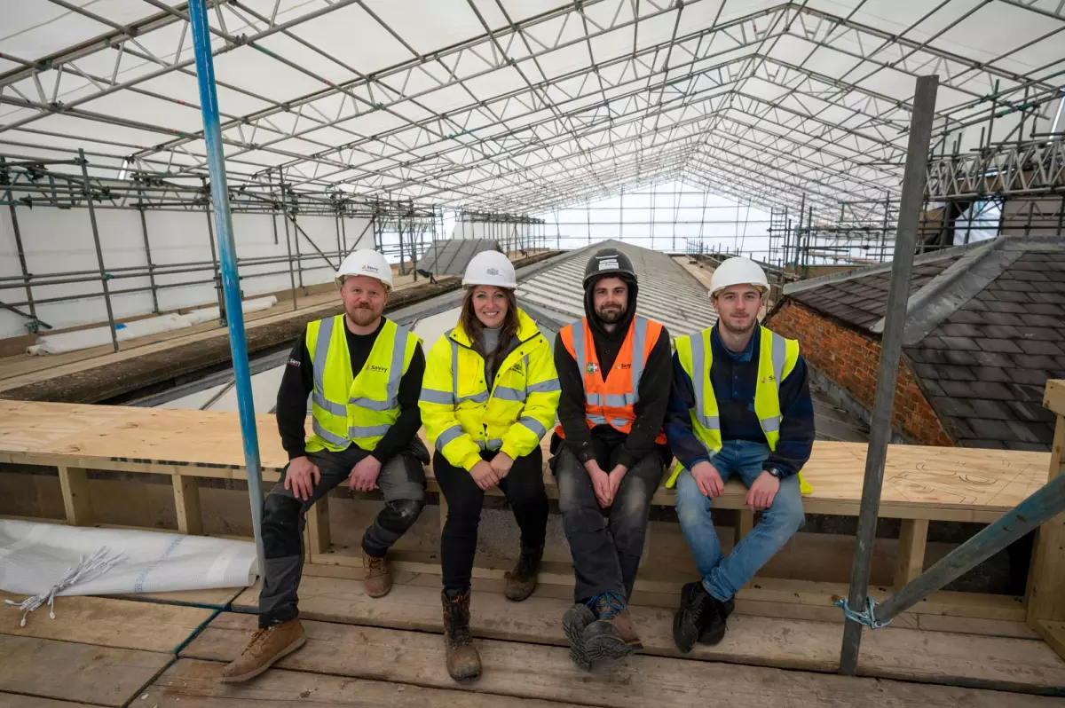 Four people in hi-vis jackets and hard hats sit on a platform beneath a scaffolding canopy during roof restoration works