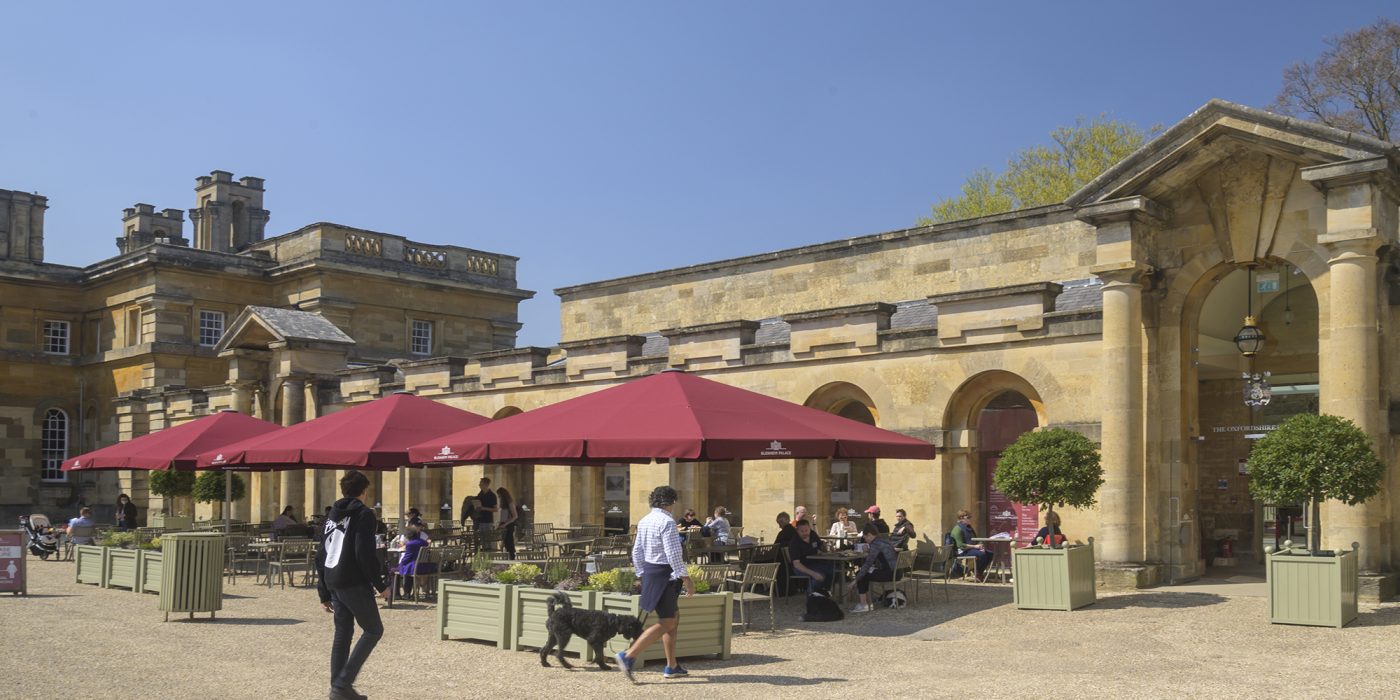 Visitors at outdoor tables under red parasols in the courtyard beside the Palace stable buildings