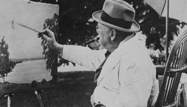 A photograph of Sir Winston Churchill painting at an easel out of doors, in a pale suit and hat