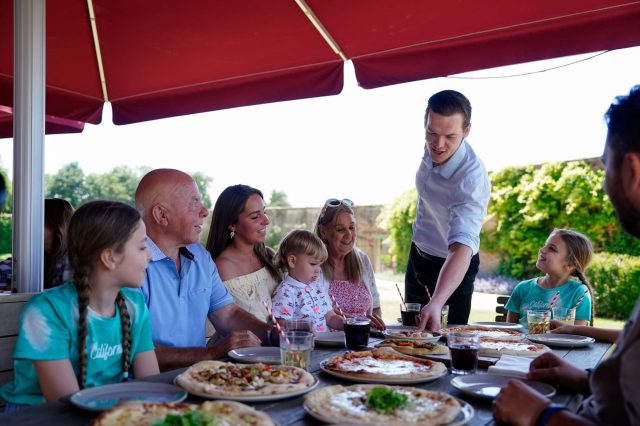 A family being served pizzas at an outdoor table under red parasols