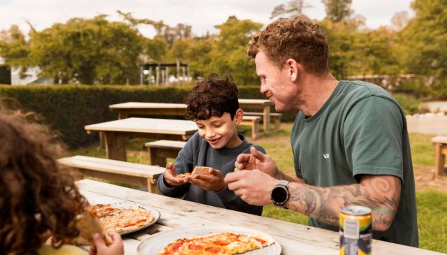 A server bringing pizzas to a family seated at a long outdoor table under a red awning