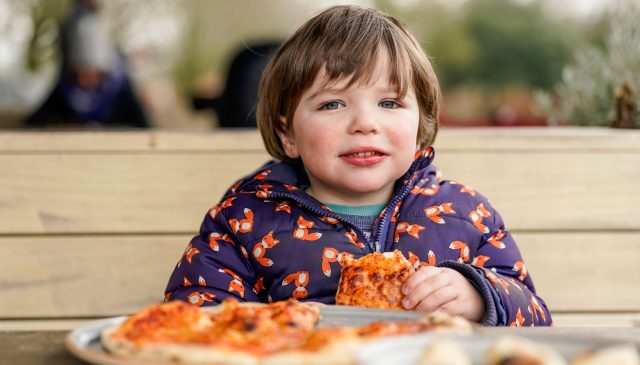 A young child in a fox-print coat eating pizza at an outdoor wooden table