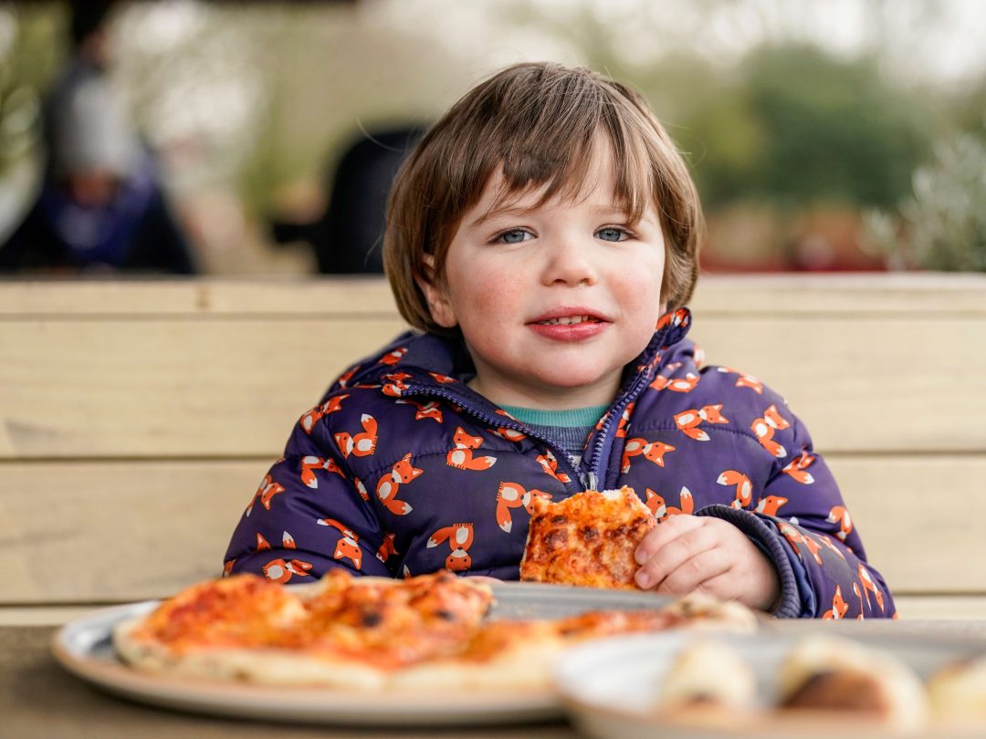 A young child in a fox-print coat eating pizza at an outdoor wooden table