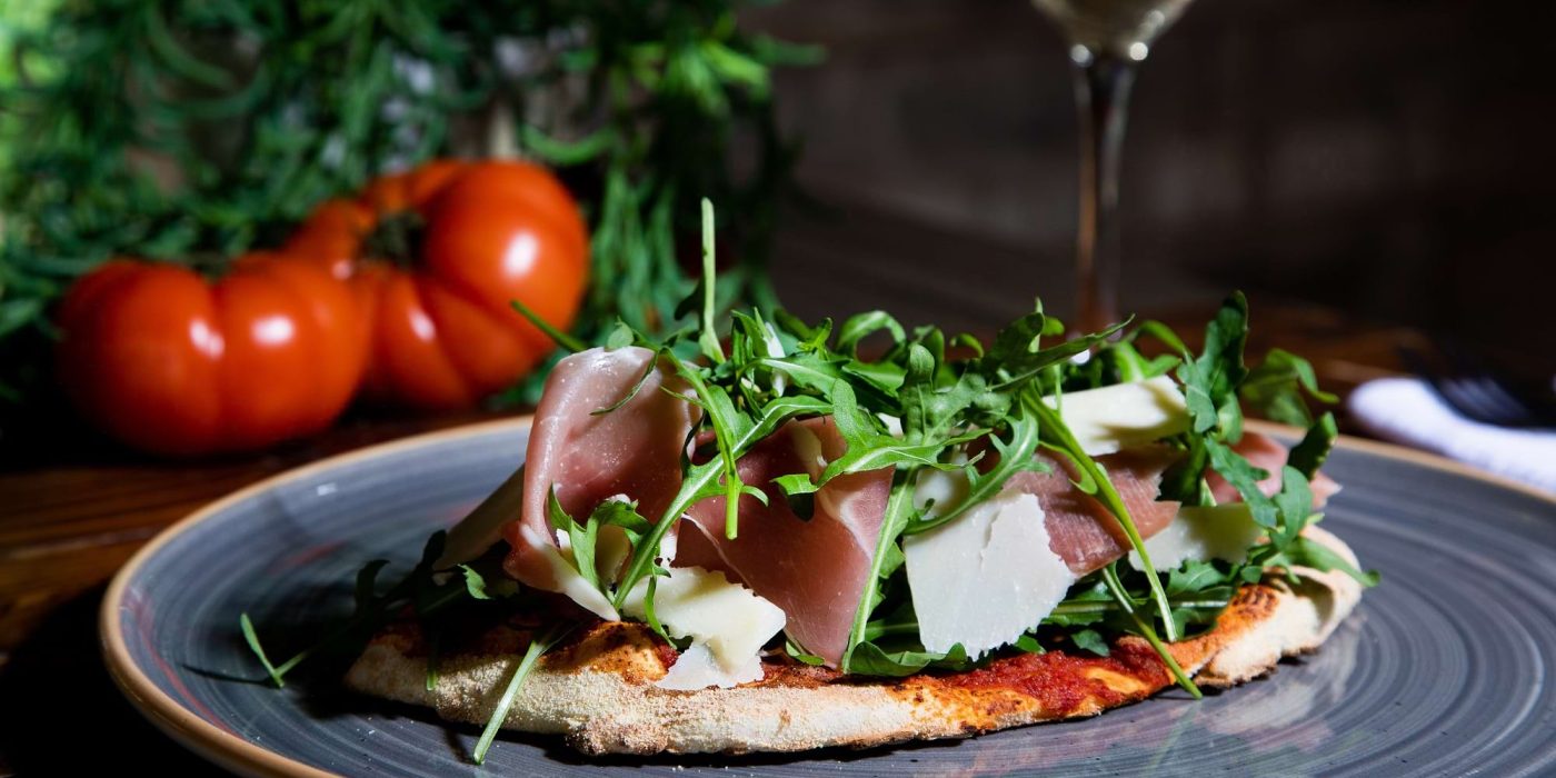 A pizza topped with cured ham, rocket and shaved cheese, with a glass of white wine