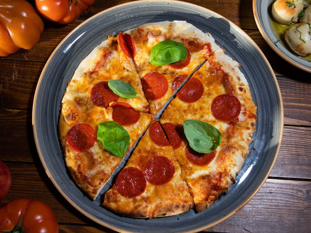 A pepperoni pizza with basil on a dark plate, with a bowl of dough balls alongside