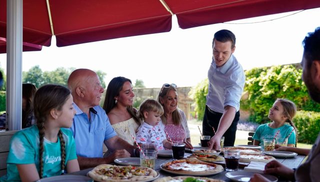 A server bringing pizzas to a family seated at a long outdoor table under a red awning