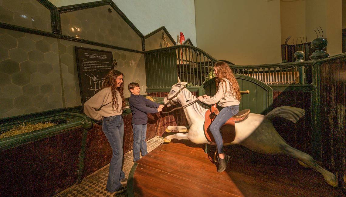 Children with a model horse in the stables exhibition, one of them seated in the saddle