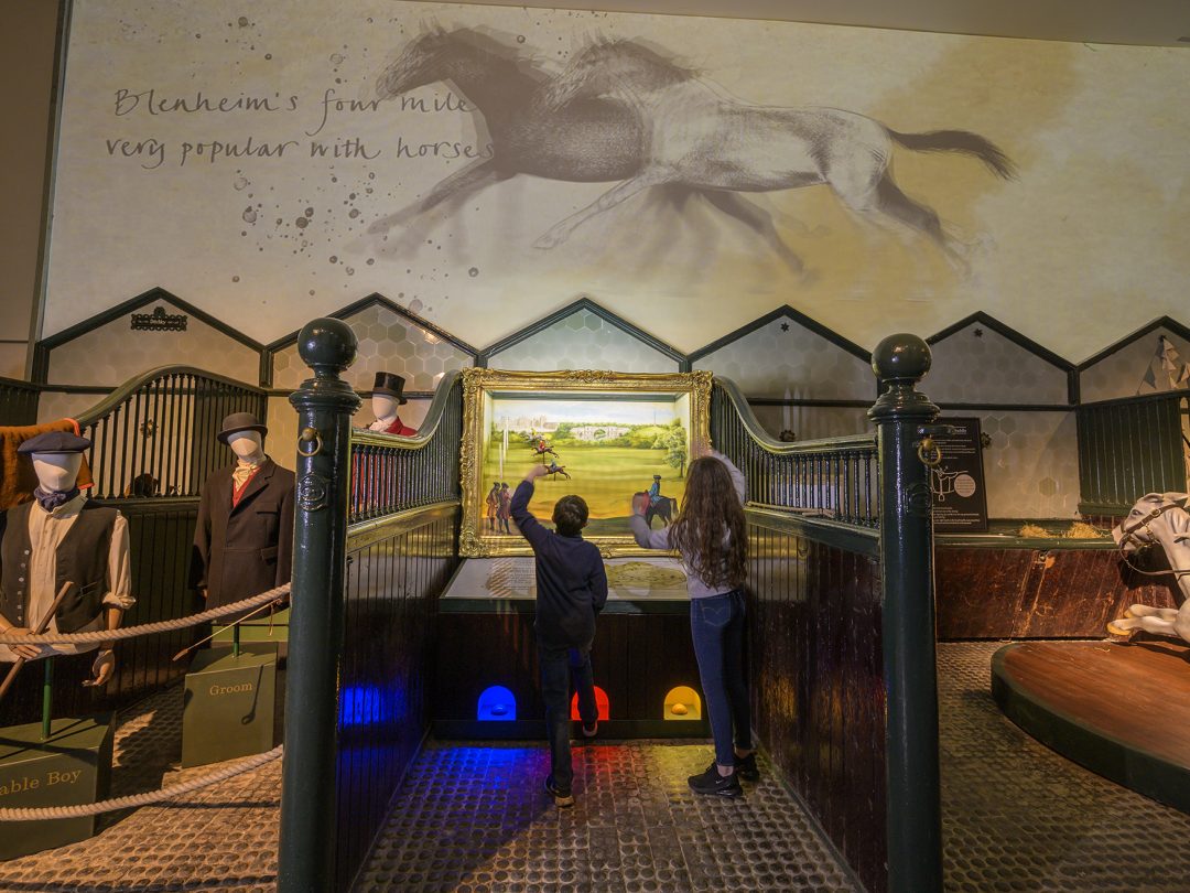 Two children at an interactive display in the stables exhibition, beside a painted horse mural