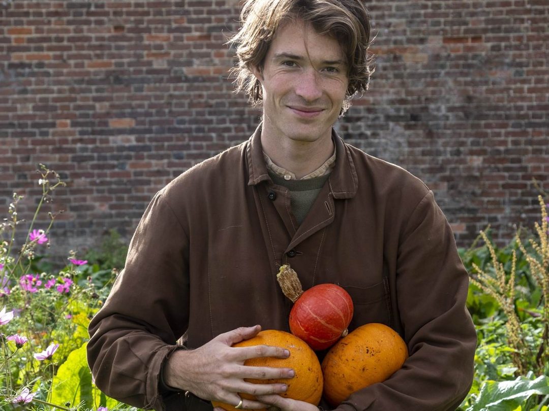 A smiling gardener in a brown work jacket holding three pumpkins in front of the brick wall of the Walled Garden