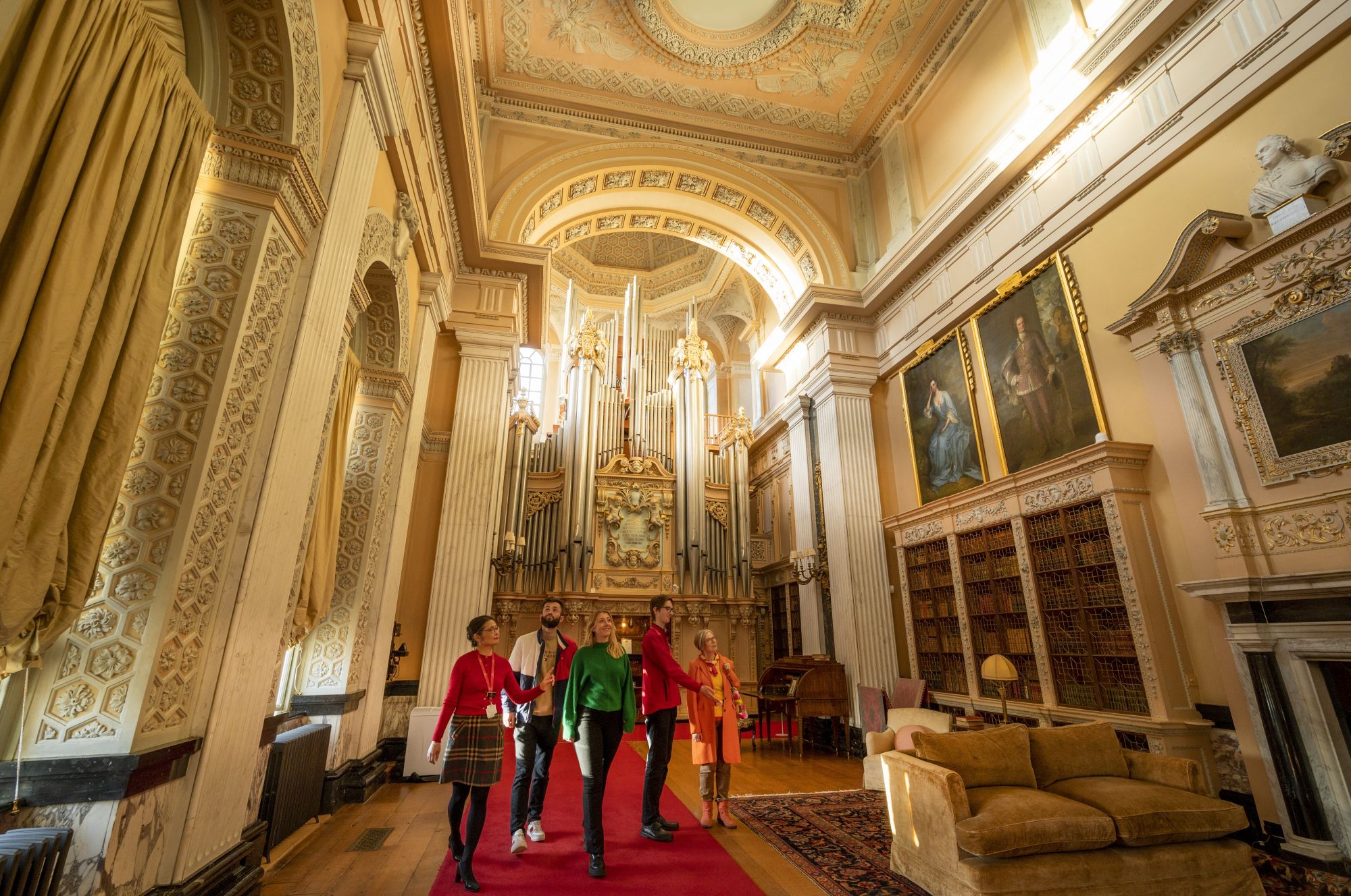 Five visitors walk along a red carpet through a long gilded library towards a grand pipe organ