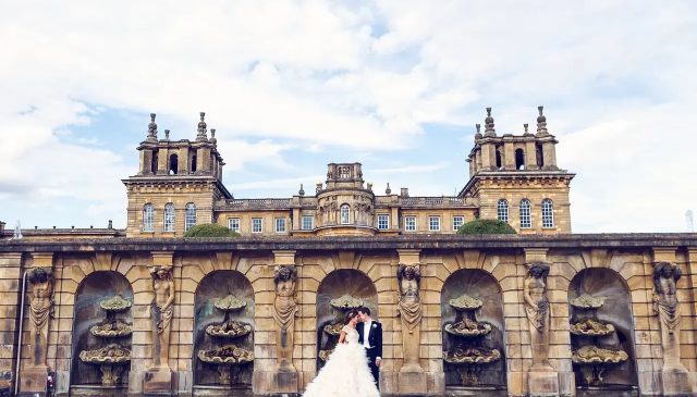 A bride and groom kiss beneath an arched stone wall with fountain niches, with the Palace rising behind