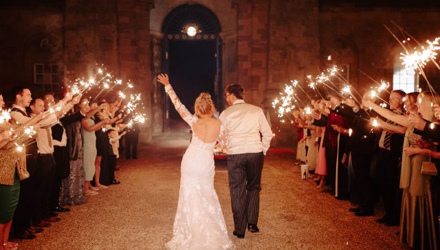 A bride and groom walk between lines of guests holding sparklers at night