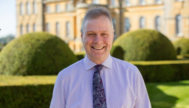 A smiling man in a shirt and patterned tie in the gardens, with clipped hedges and the Palace