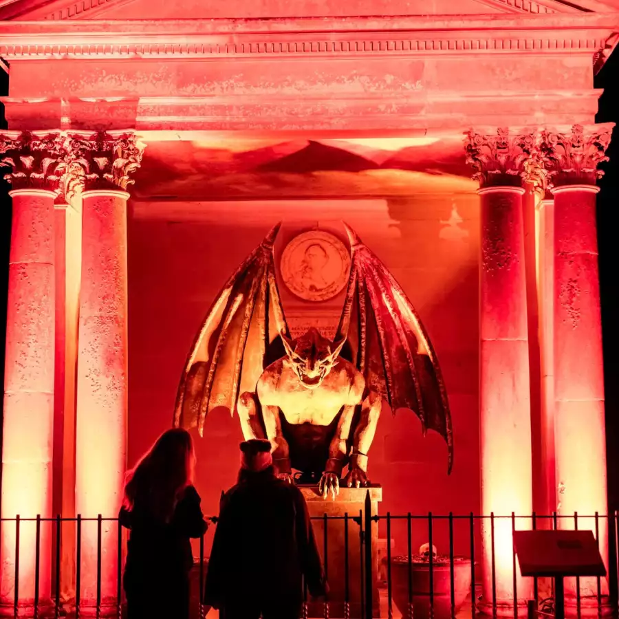 Winged gargoyle figure lit red inside a classical temple, watched by two visitors