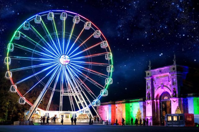 A brightly lit observation wheel at night beside the illuminated Palace gateway