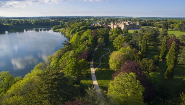 Aerial view of the lake and tree-lined parkland in spring, with the Palace on the far side beneath a blue sky
