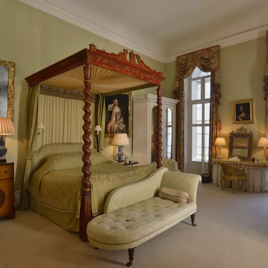 A carved four-poster bed and a pale green chaise longue in a high-ceilinged bedroom with portraits