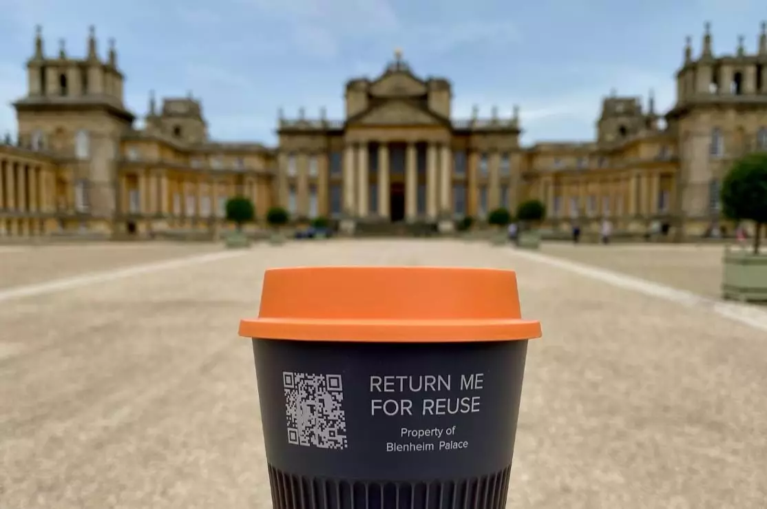 A dark reusable coffee cup with an orange lid marked Return me for reuse, held up by the Palace