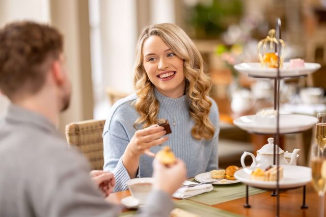 A couple enjoy a festive afternoon tea by a Christmas tree, the woman wearing a tiara